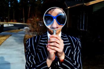 person wearing sunglasses and a striped black suit holding a magnifying glass up to their face